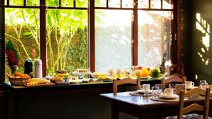 Une table mise pour le petit-déjeuner, avec une place pour deux personnes, une vue sur la verdure par la fenêtre, et une variété d'aliments proposés, notamment des fruits, des viennoiseries et des boissons.