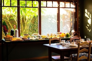 A breakfast setting with a table set for two, a window view of greenery, and a variety of breakfast items including fruits, pastries, and beverages. - Bollté Praia Boutique Pousada - Campeche - SC (Florianópolis)