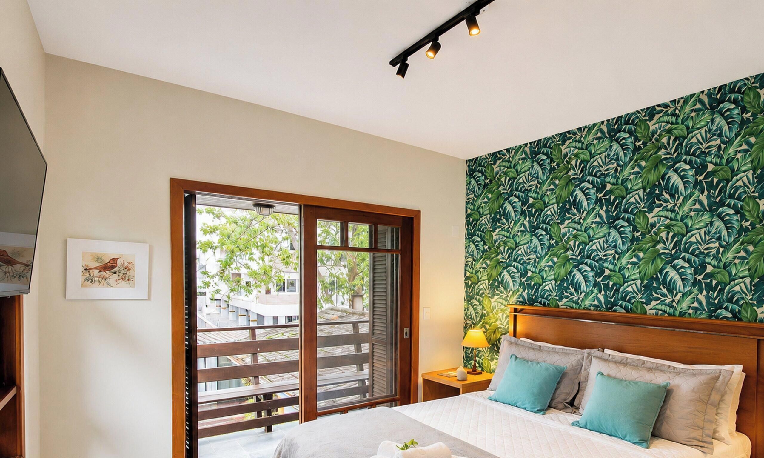 A bedroom with a bed, a nightstand, a lamp, a window with a view, and a wall with a leaf pattern.