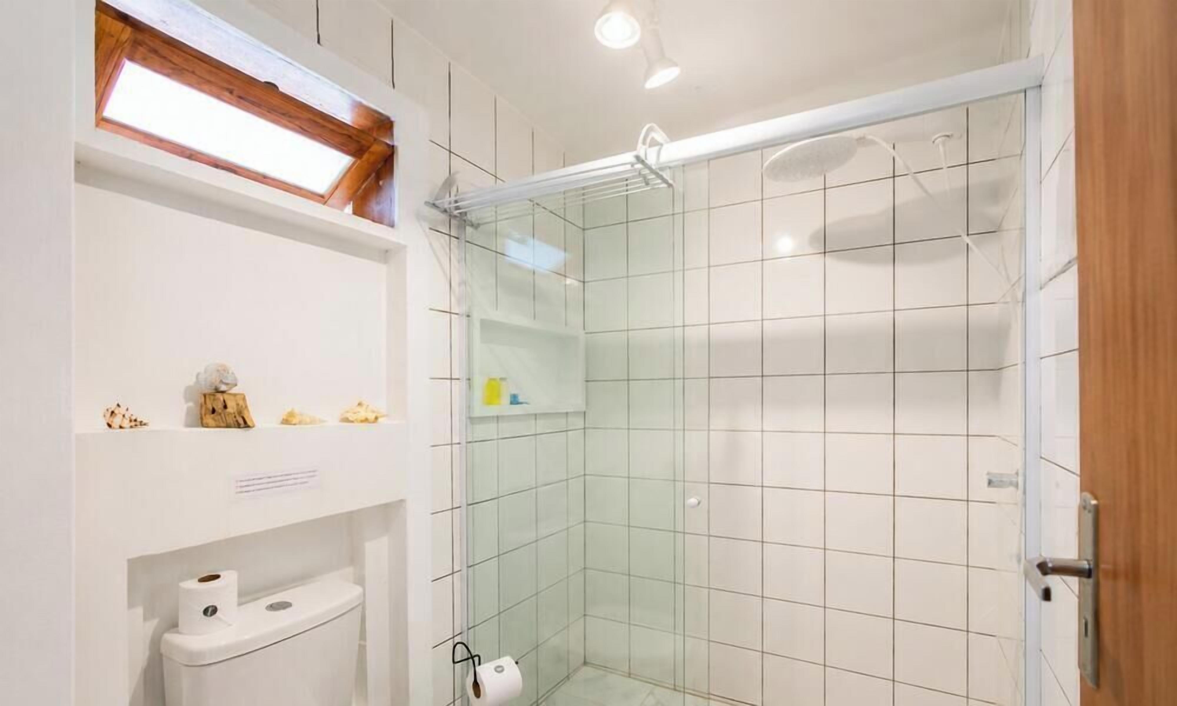 A modern bathroom with a glass shower enclosure, white tiles, a toilet, and a shelf with toiletries.