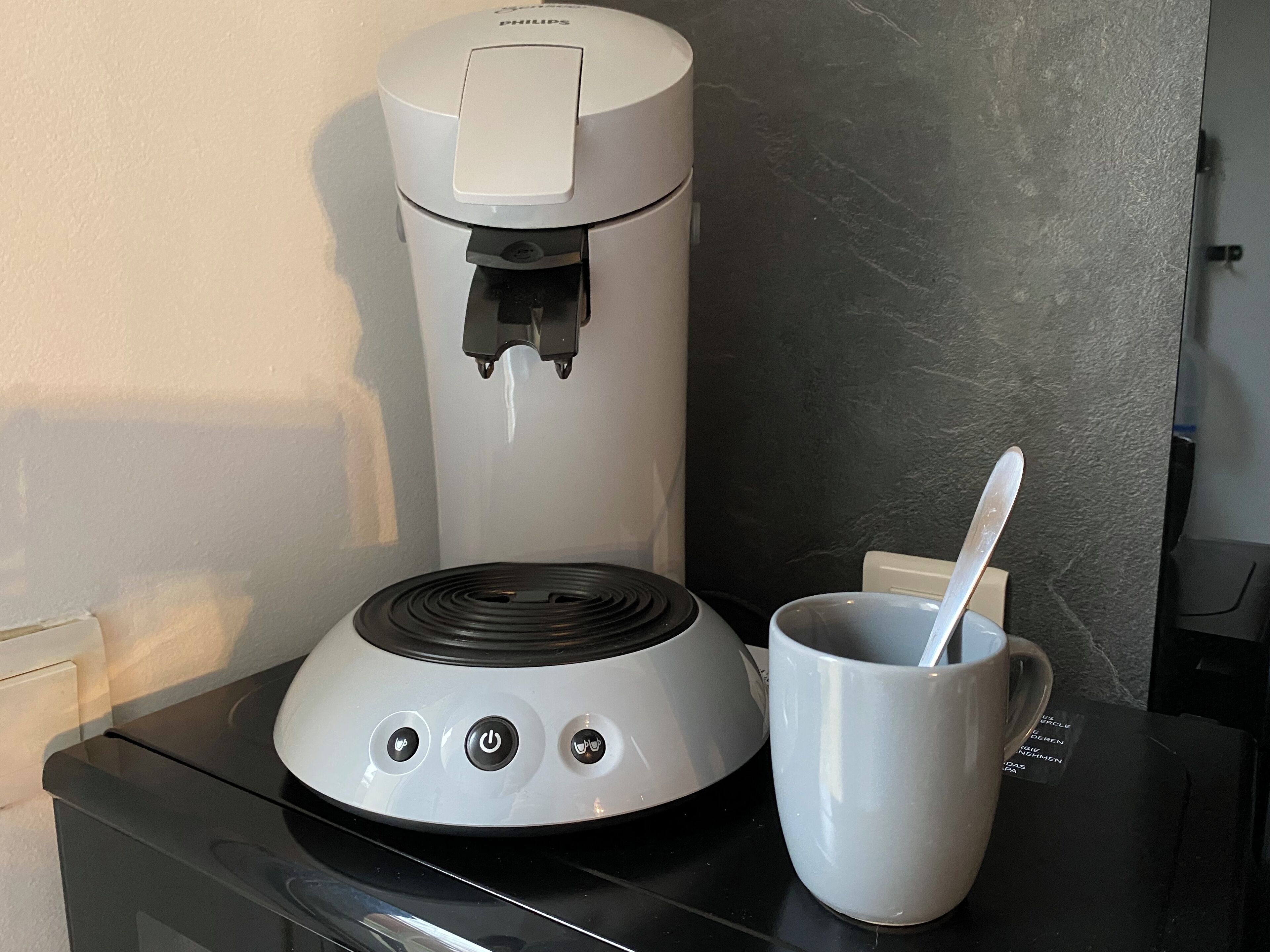 Eine Kaffeemaschine mit Tasse und Löffel auf einer schwarzen Arbeitsplatte.