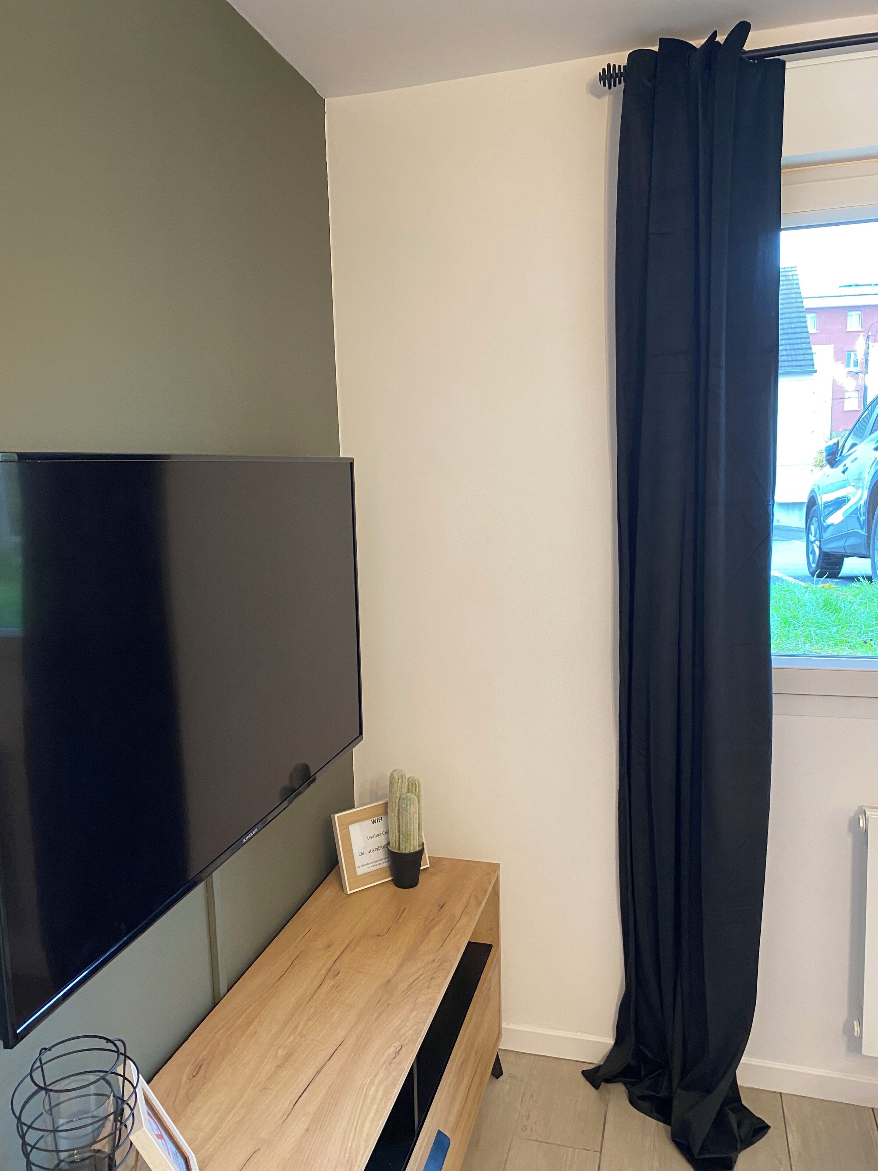 A modern living room with a flat-screen TV mounted on the wall, a wooden coffee table with a cactus, and a black curtain covering a window.
