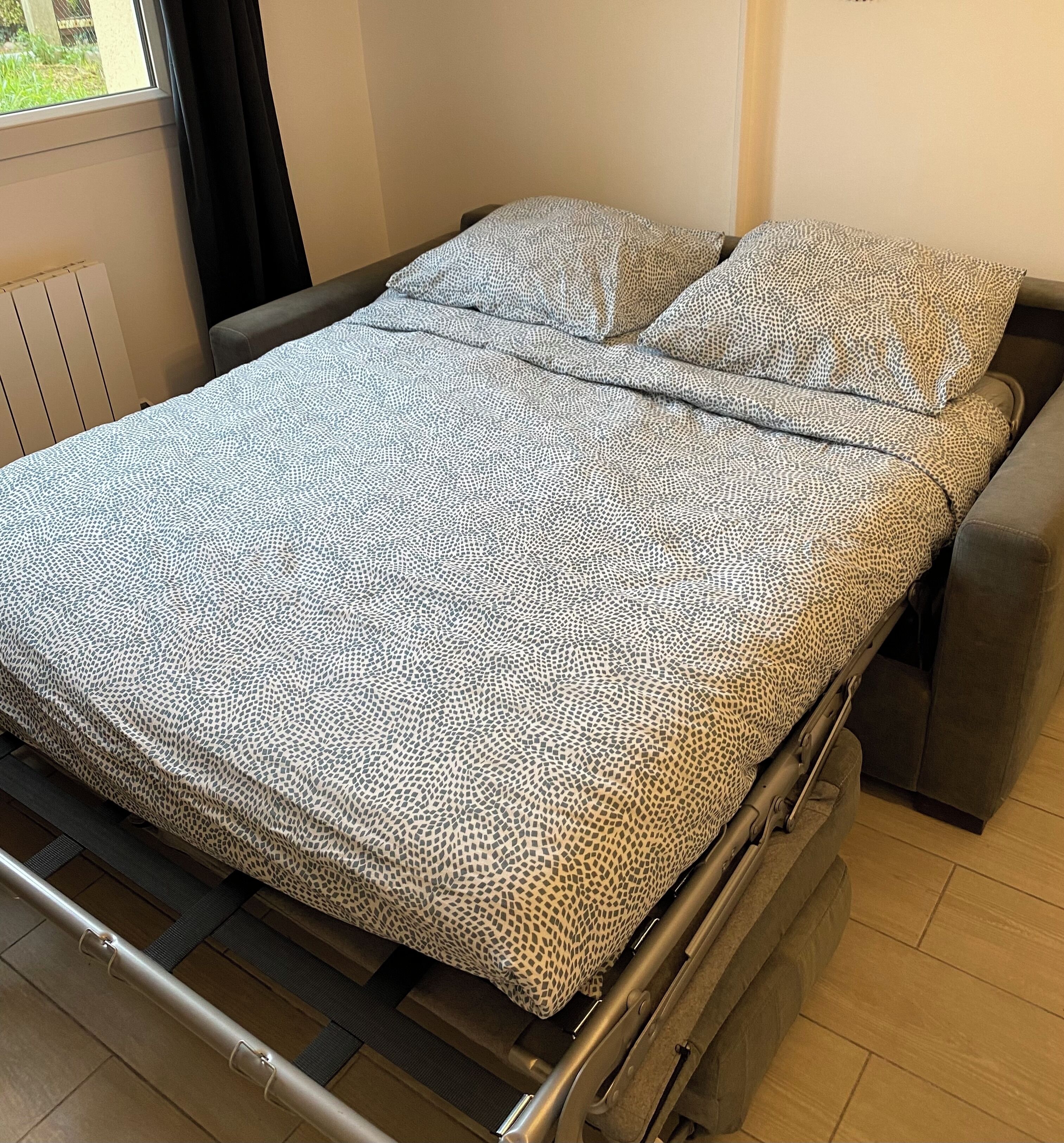 A hospital bed with a patterned blanket and pillows, a window with curtains, and a radiator on the wall.