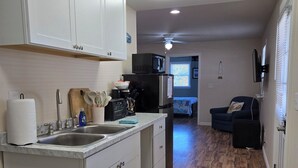 Una cocina moderna con armarios blancos, encimera de mármol y microondas integrado. El piso es de madera, hay un sofá oscuro en la sala de estar y un ventilador de techo.