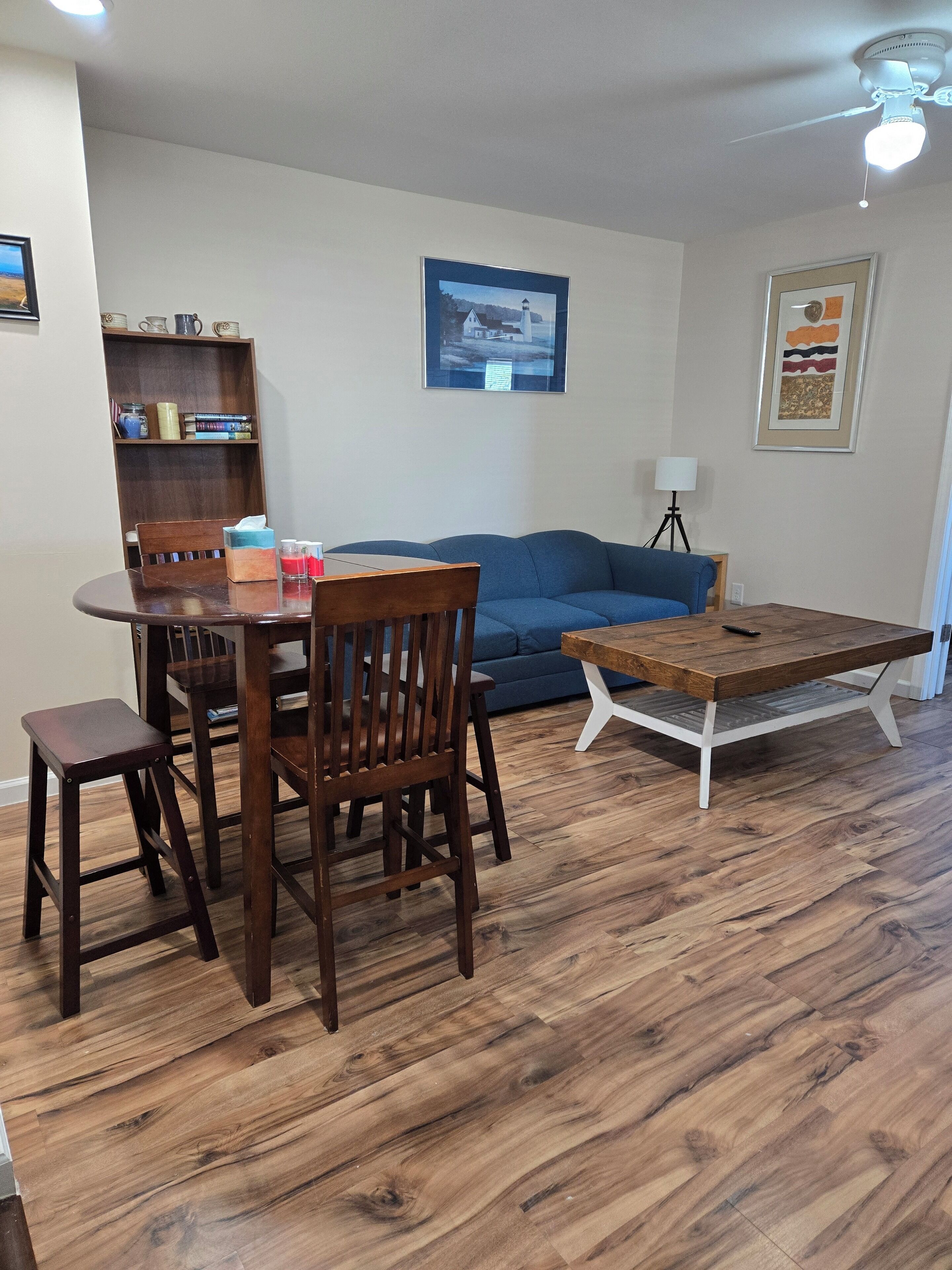 Una sala de estar con una mesa de comedor de madera, sillas, un sofá azul, una estantería y un cuadro enmarcado en la pared.