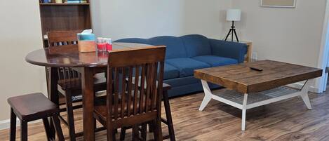 Una sala de estar con una mesa de comedor de madera, sillas, un sofá azul, una estantería y un cuadro enmarcado en la pared.