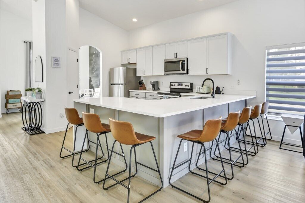 Een moderne keuken met witte kasten, een groot eiland met barkrukken, roestvrijstalen apparatuur en een raam met jaloezieën.