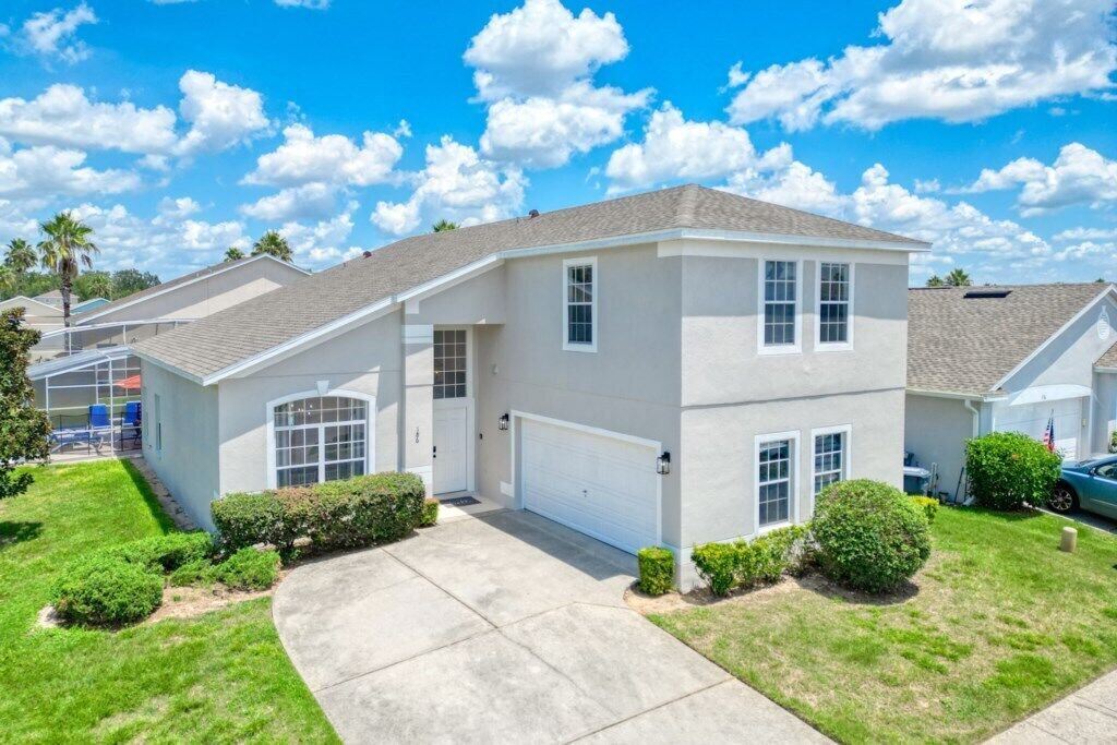 A two-story residential house with a garage, a front yard with a lawn and bushes, and a clear blue sky with clouds.