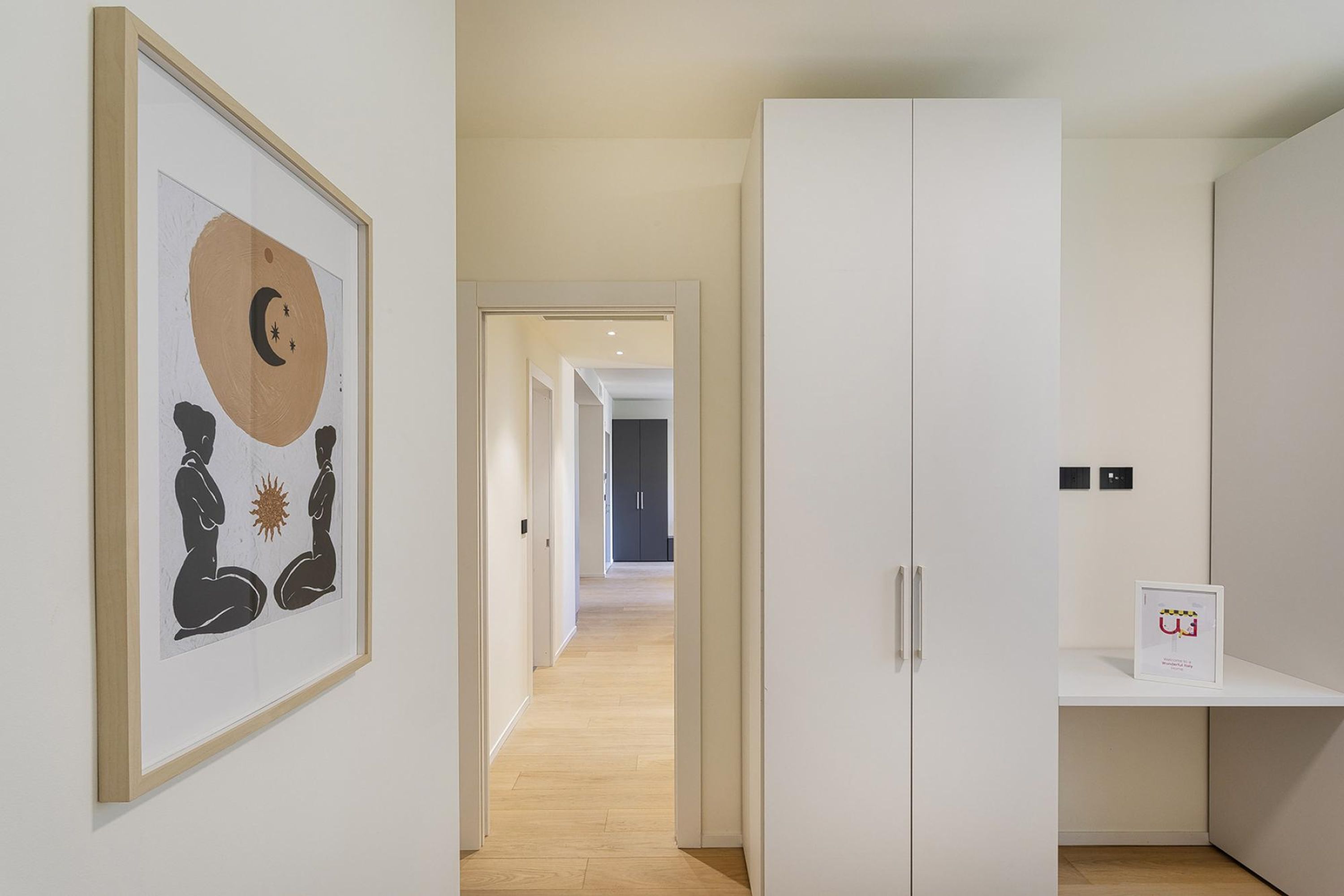 A hallway with a framed artwork, a white wardrobe, and a small shelf with a framed item.