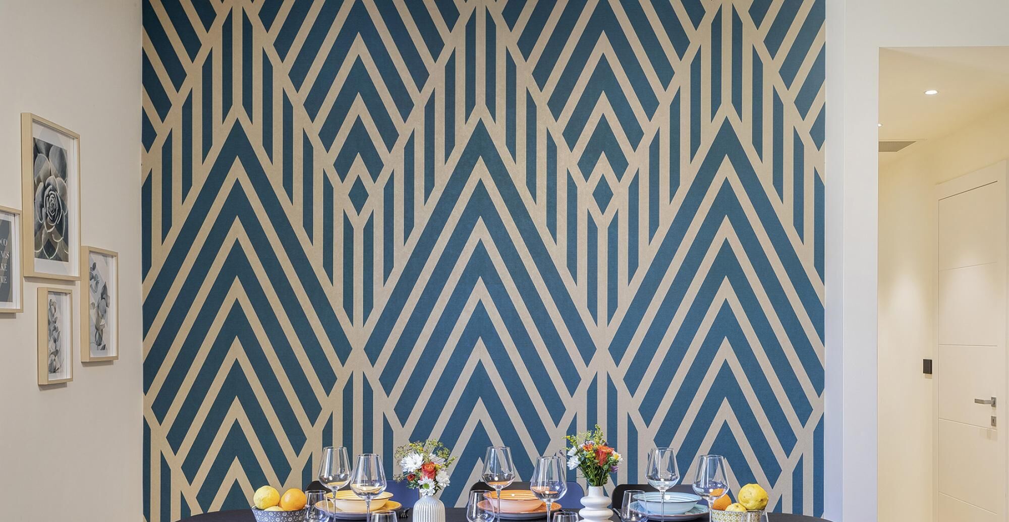 Un comedor con una pared de estampado geométrico, una mesa preparada para cuatro personas, con platos, vasos y un florero.