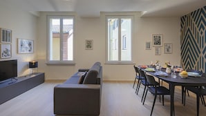 Moderni olohuone, jossa on sohva, ruokapöytä ja tuolit. Huoneessa on suuret ikkunat, taideteoksia seinillä ja litteänäyttöinen televisio jalustalla.