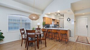 Een moderne keuken met houten kastjes, een centraal eiland met barkrukken, een zithoek met een ronde tafel en stoelen, en een groot raam met jaloezieën.