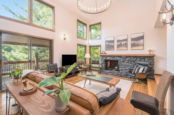 Un salon spacieux avec une cheminée en pierre, des canapés en cuir, une table basse et un téléviseur. De grandes fenêtres offrent une vue sur la verdure.