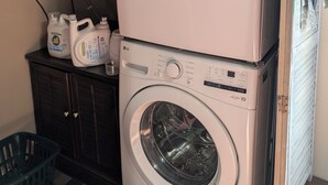 Een gestapelde wasmachine en droger met een bedieningspaneel, een plank erboven met schoonmaakmiddelen en een wasmand eronder.