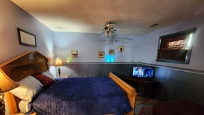 Een slaapkamer met een bed, nachtkastje, lamp, plafondventilator, televisie en een raam met jaloezieën.