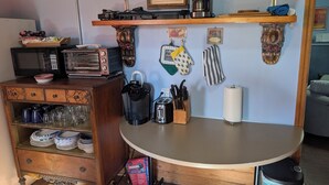 Een keuken met een magnetron, broodrooster, waterkoker en koffiezetapparaat op het aanrecht. Boven het aanrecht staan planken met een lijst, een koffiekan en een opgerolde handdoek. Een prullenbak is zichtbaar onder het aanrecht.