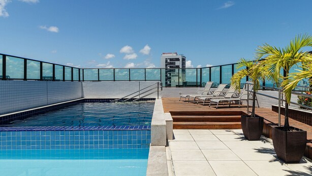 A rooftop pool with clear blue water, surrounded by a glass railing, and a wooden deck with lounge chairs and potted plants. - Hy Beach Flats - Santa Maria (Recife)