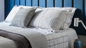 A neatly made bed with a quilted white comforter and two pillows, a blue headboard, and a bedside table with a lamp and a decorative item. - Hy Beach Flats - Santa Maria (Recife)