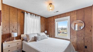 Een slaapkamer met houten paneelwanden, een bed met een witte sprei, een nachtkastje met een lamp, een raam met uitzicht, en een ronde spiegel aan de muur.