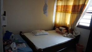 Un lit simple avec un cadre en bois, un matelas blanc et deux oreillers. On aperçoit une petite table recouverte d’une nappe bleue, sur laquelle sont posés divers objets. Une fenêtre avec des rideaux est visible à l’arrière-plan.