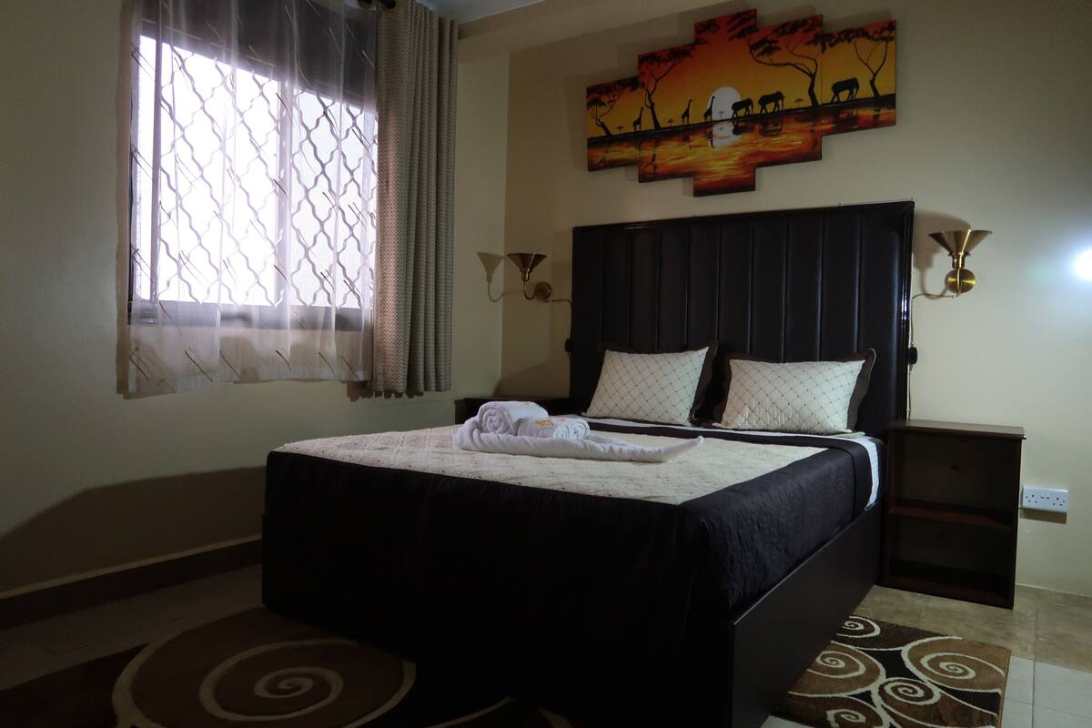 A neatly made bed with a dark headboard, a wall-mounted artwork depicting a sunset, and a window with a patterned curtain.
