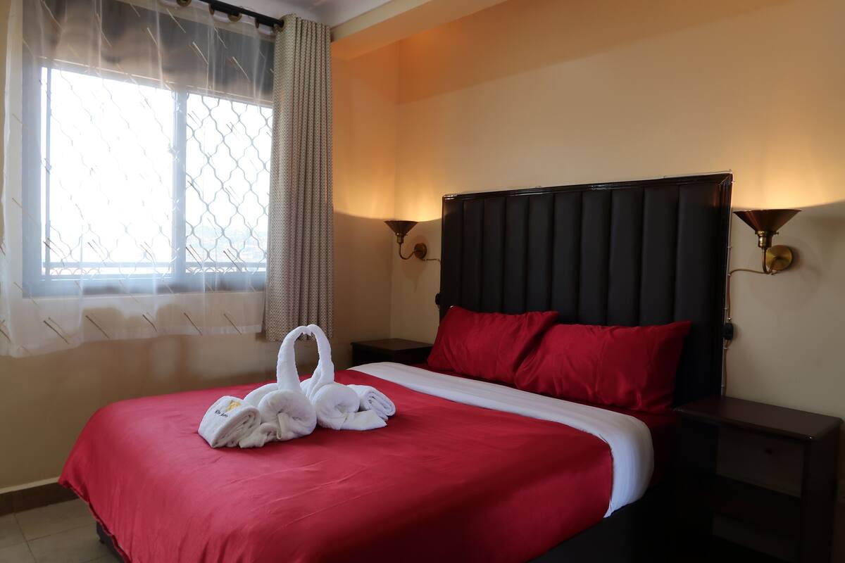 A hotel room with a bed, red bedding, two red pillows, a black headboard, and two wall-mounted lamps.