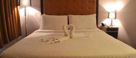 A hotel room with a large bed, two pillows, a headboard, a nightstand with a lamp, and a swan-shaped towel on the bed.