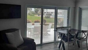 A living room with a dark-colored sofa, a glass-top dining table with chairs, and a large sliding glass door leading to an outdoor patio area. - Sea Biscuit Ocean Villa 911 (Abaco)