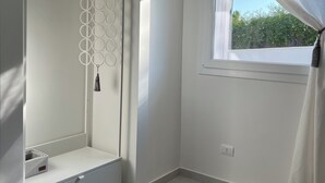 Une chambre à coucher moderne et minimaliste, dotée d’une armoire intégrée, d’une petite fenêtre avec des rideaux légers et offrant une vue sur la verdure environnante.