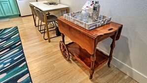 Een houten karretje met een dienblad waarop een cocktailshaker, glazen en een vaas met bloemen staan, geplaatst in een ruimte met een keuken op de achtergrond.