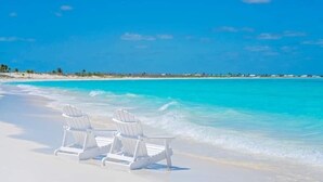 Deux chaises de plage blanches sur une plage de sable, avec une eau turquoise cristalline et un ciel bleu dégagé.