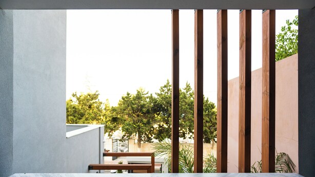 Un área de estar moderna al aire libre con una mesa y sillas de madera, una pared de concreto y vistas a árboles y un edificio al fondo.