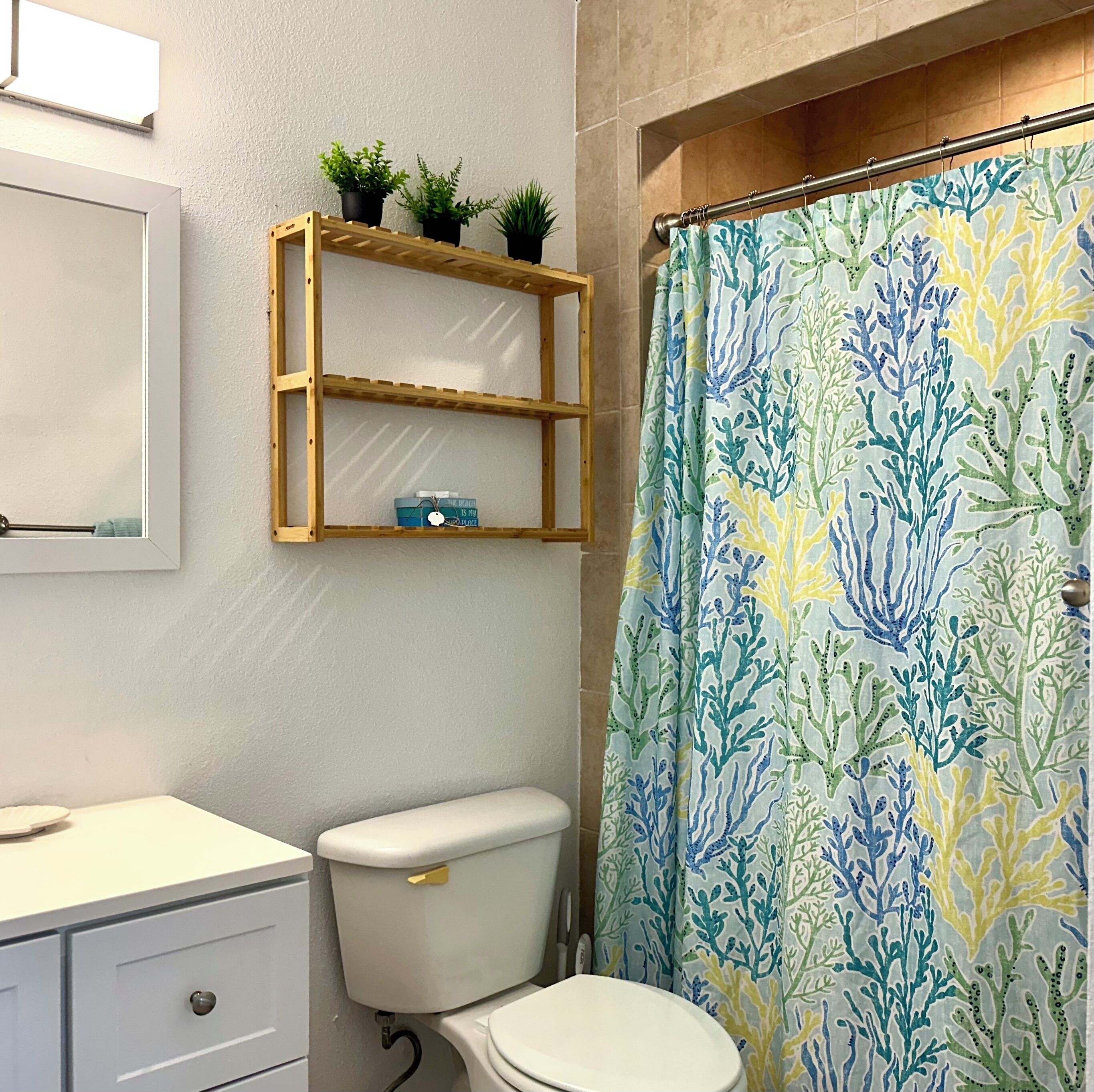 A bathroom with a toilet, a wooden shelf with plants, and a shower curtain with a coral pattern.