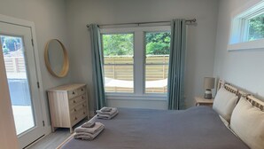 A bedroom with a large bed, a wooden dresser, a round mirror, and a window with curtains. - Florida style  built  2023. Heated pool. Dog park. 10 min walk to beach.  (Southern Shores)