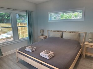 Cama bem arrumada com duas toalhas dobradas, mesa de cabeceira com abajur e janela com cortinas.
