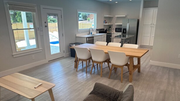 A modern kitchen with white cabinets, a stainless steel refrigerator, and a dining area with a wooden table and white chairs. - Florida style  built  2023. Heated pool. Dog park. 10 min walk to beach.  (Southern Shores)