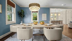 A dining room with a large table set for six, surrounded by six chairs. There is a chandelier above the table, and a potted plant to the left.