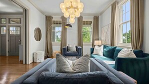 A spacious living room with a large sofa, a smaller couch, and a chair. There is a chandelier hanging from the ceiling, and large windows with curtains.