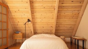 Une chambre avec un plafond en pente, un plancher en bois, un lit, une table de chevet avec une lampe et un petit tabouret en bois.