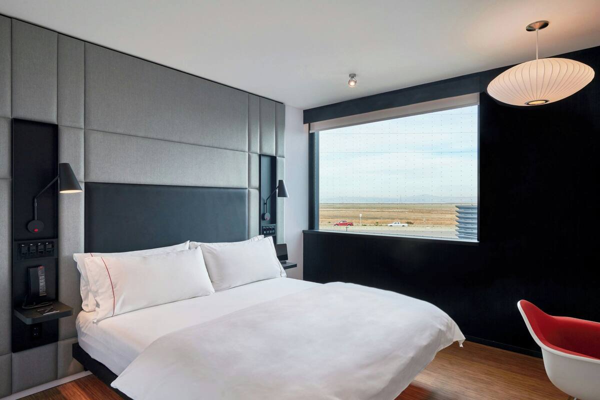 A modern hotel room with a large bed, a side table with a lamp, a red chair, and a view of the desert through a large window.