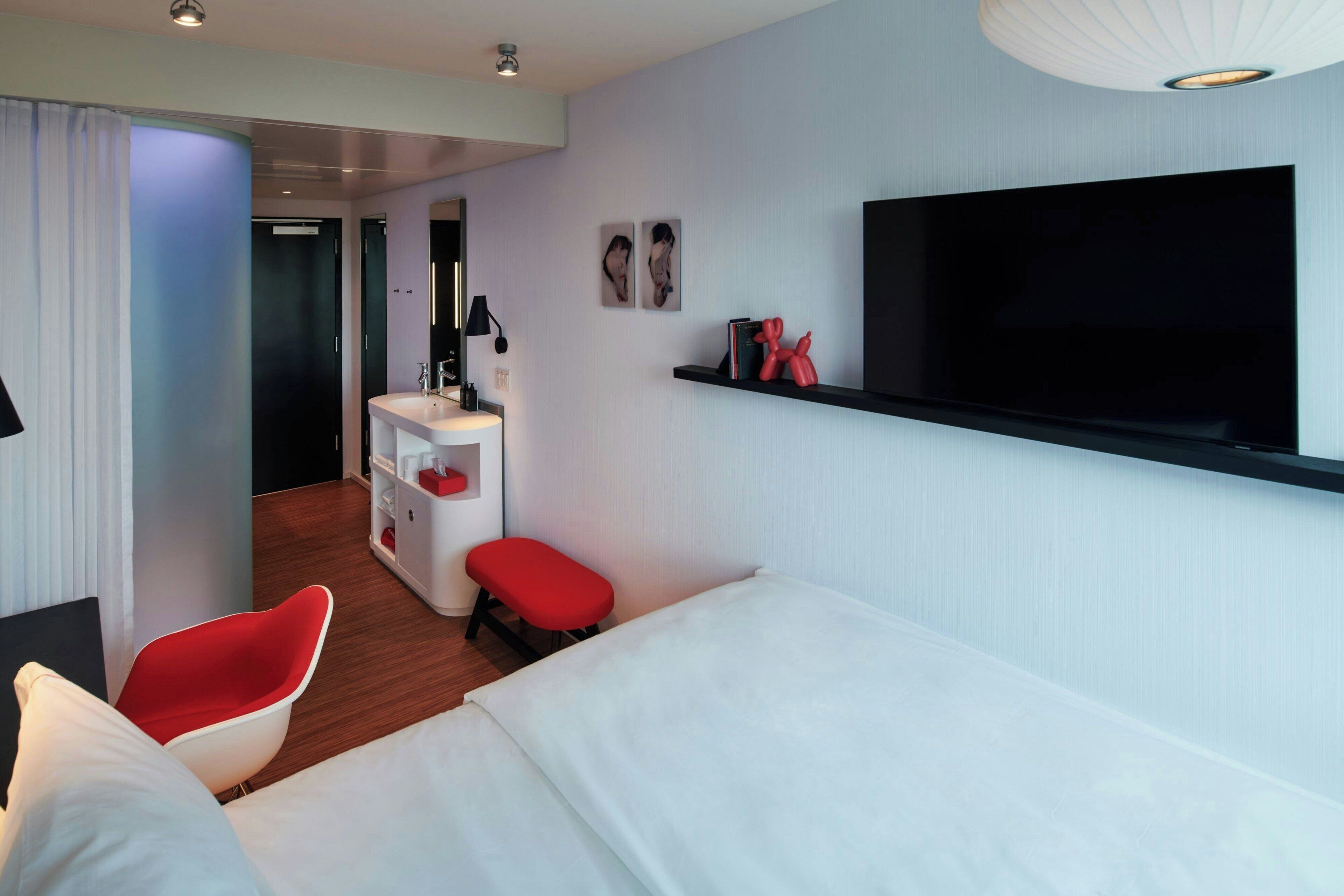 Ein modernes Hotelzimmer mit einem Bett, einem roten Sessel, einem kleinen Schreibtisch mit Spiegel, einem an der Wand befestigten Fernseher und einem Regal mit Dekoration.