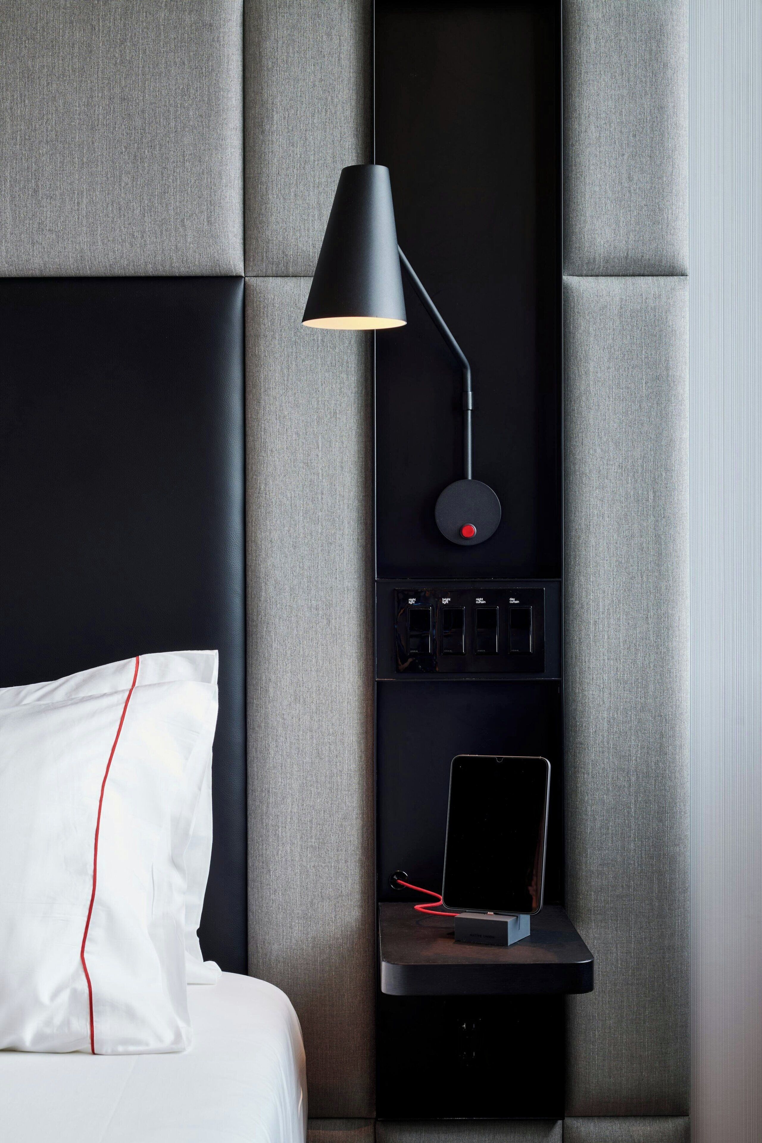 Ett modernt hotellrum med en säng, ett sängbord med en lampa, ett grenuttag och en laddningsstation.