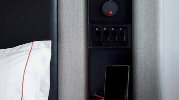Ett modernt hotellrum med en säng, ett sängbord med en lampa, ett grenuttag och en laddningsstation.