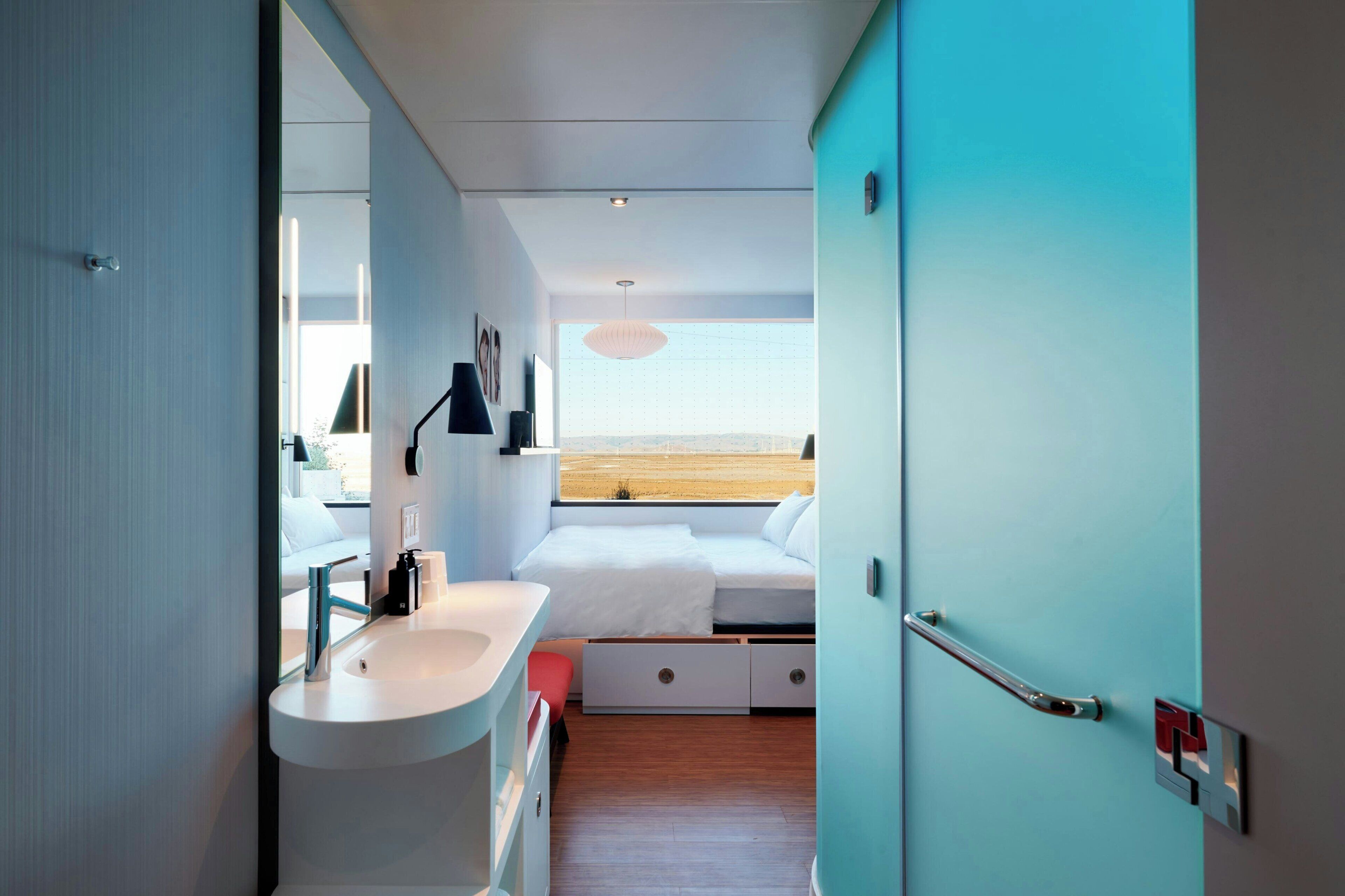 Un dormitorio moderno con un ventanal grande, una cama, una mesita de noche y un baño con lavabo y espejo.
