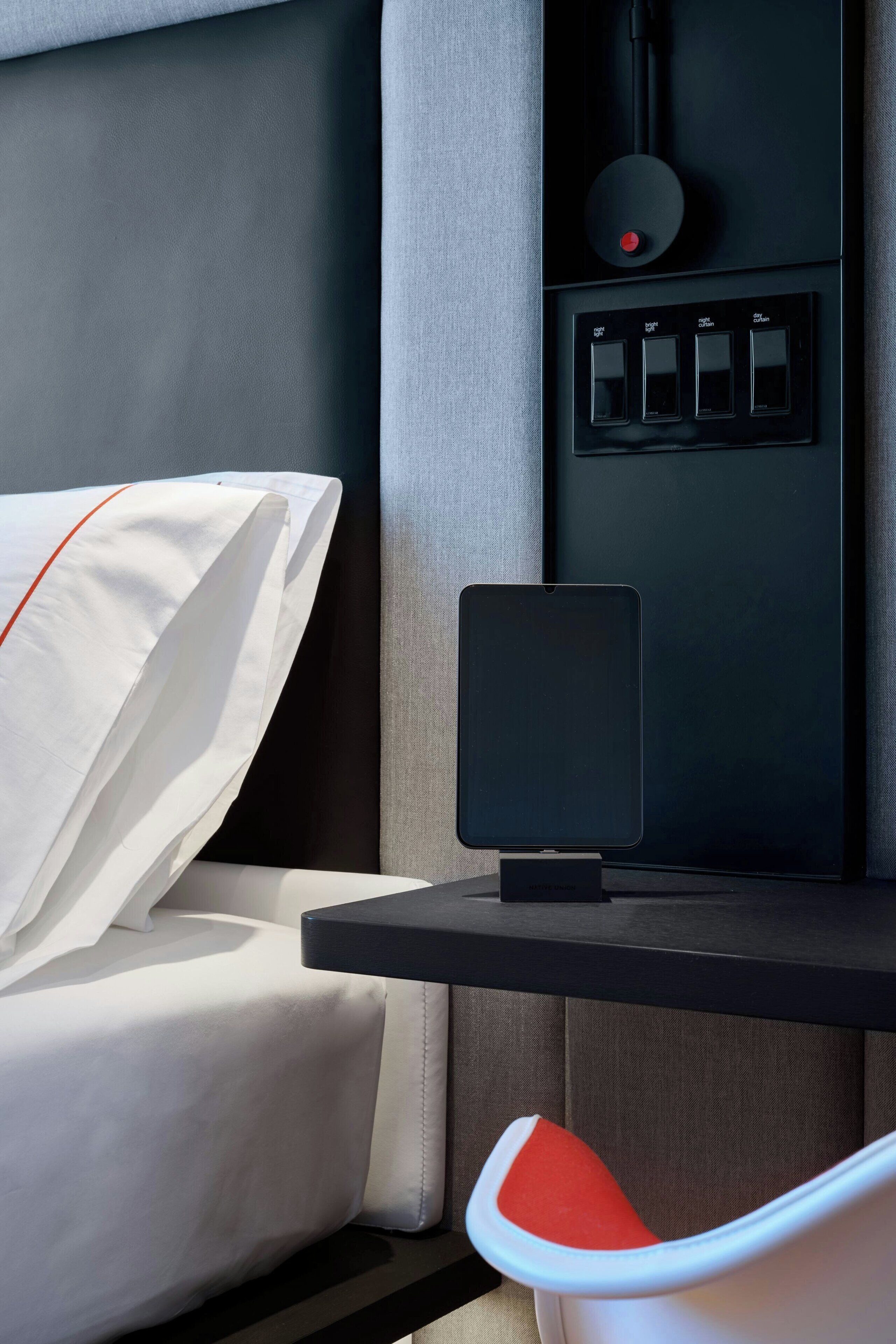 Una cama bien tendida con sábanas blancas y un detalle rojo, una mesita de noche negra con una tableta, y un panel de control con cuatro botones.
