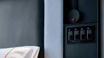 Una cama bien tendida con sábanas blancas y un detalle rojo, una mesita de noche negra con una tableta, y un panel de control con cuatro botones.
