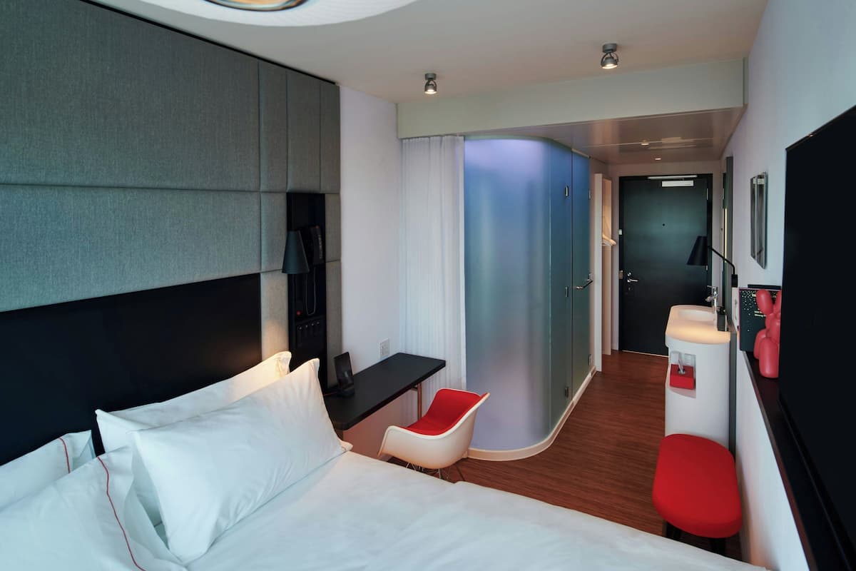 A modern hotel room with a bed, a desk, a chair, a shower area, and a sink.