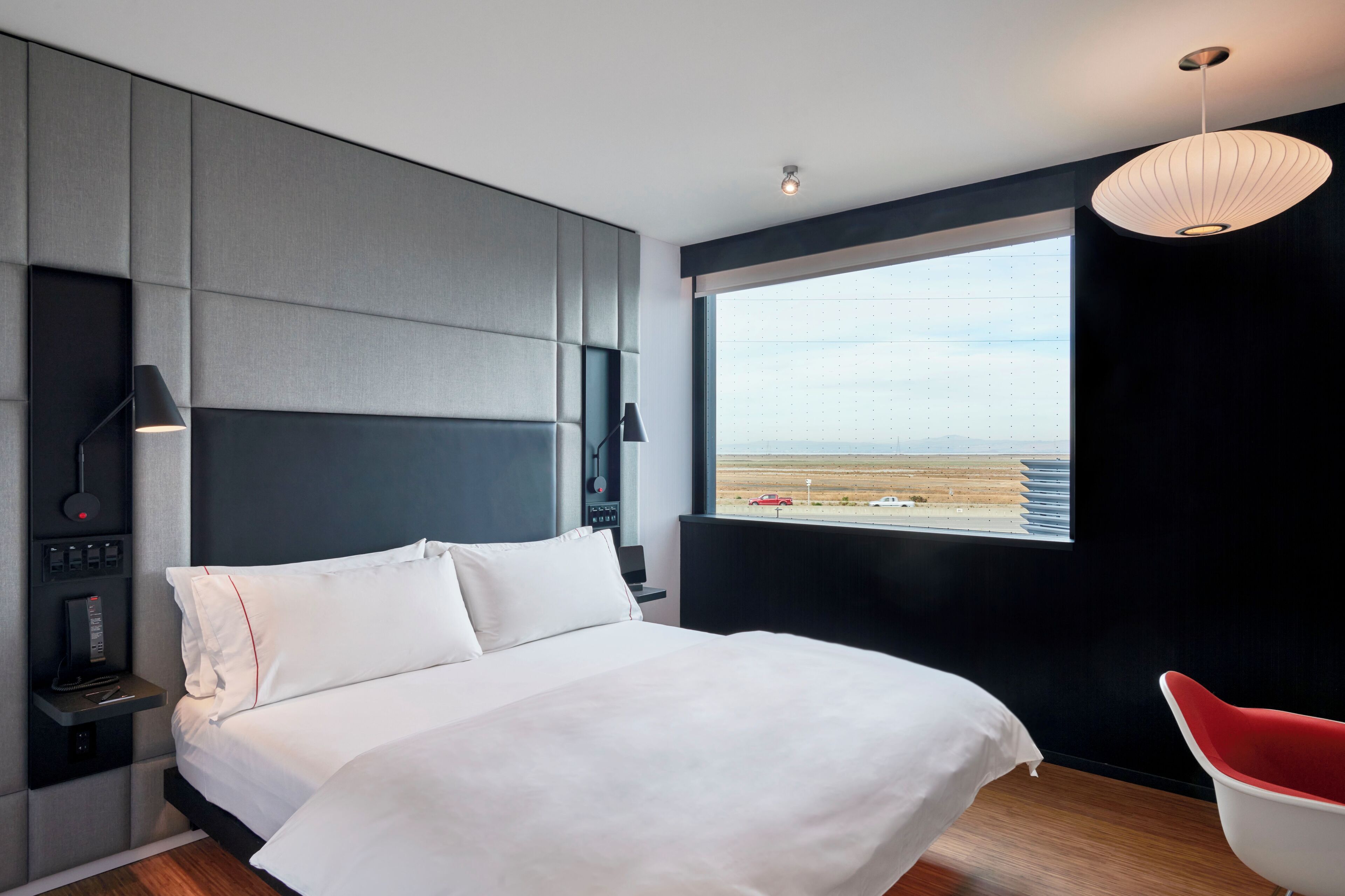 A modern hotel room with a large bed, a view of the desert, and a red chair.