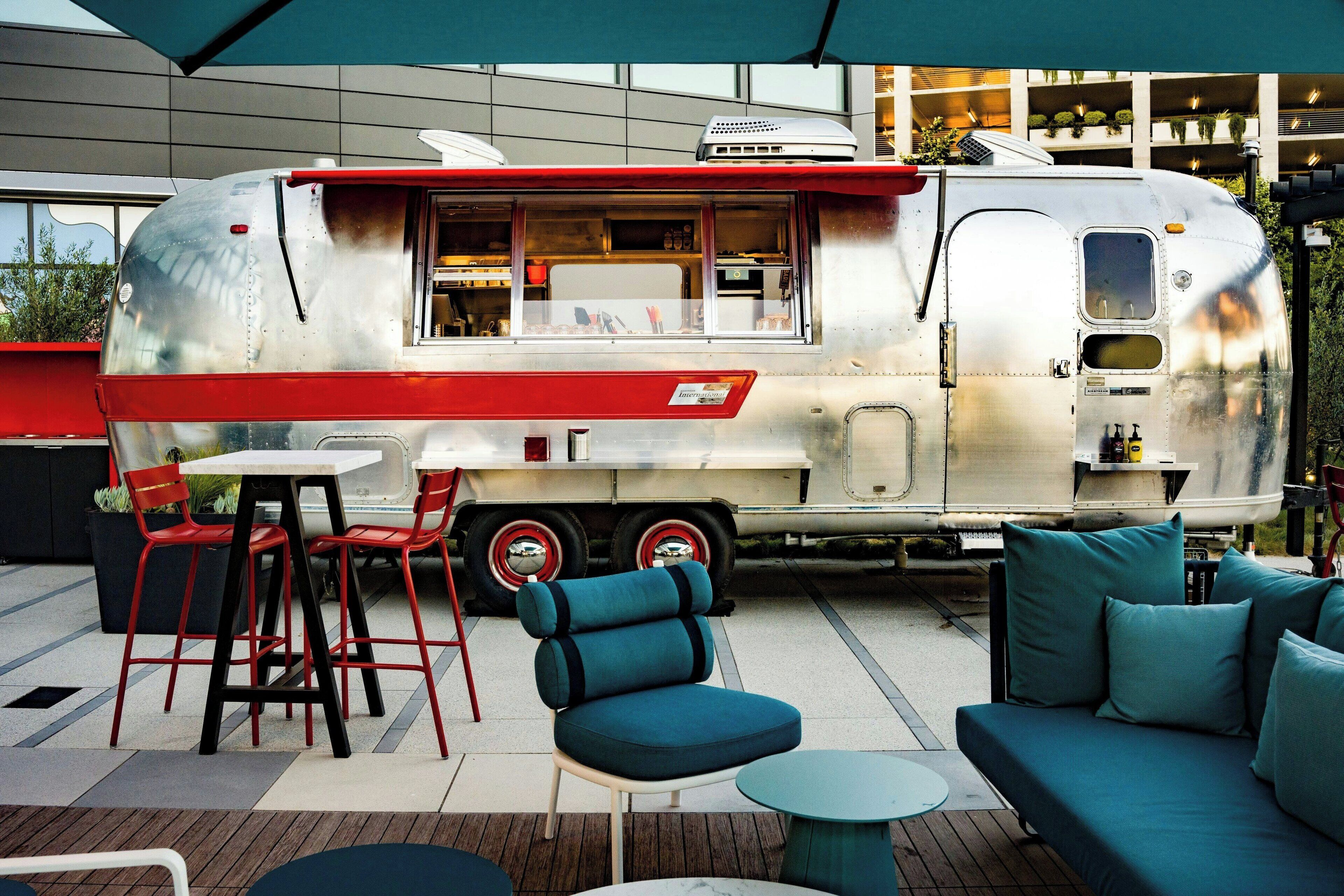 A vintage food truck with a red and white exterior parked in an outdoor seating area with blue furniture and a modern building in the background.