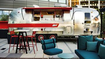 A vintage food truck with a red and white exterior parked in an outdoor seating area with blue furniture and a modern building in the background.